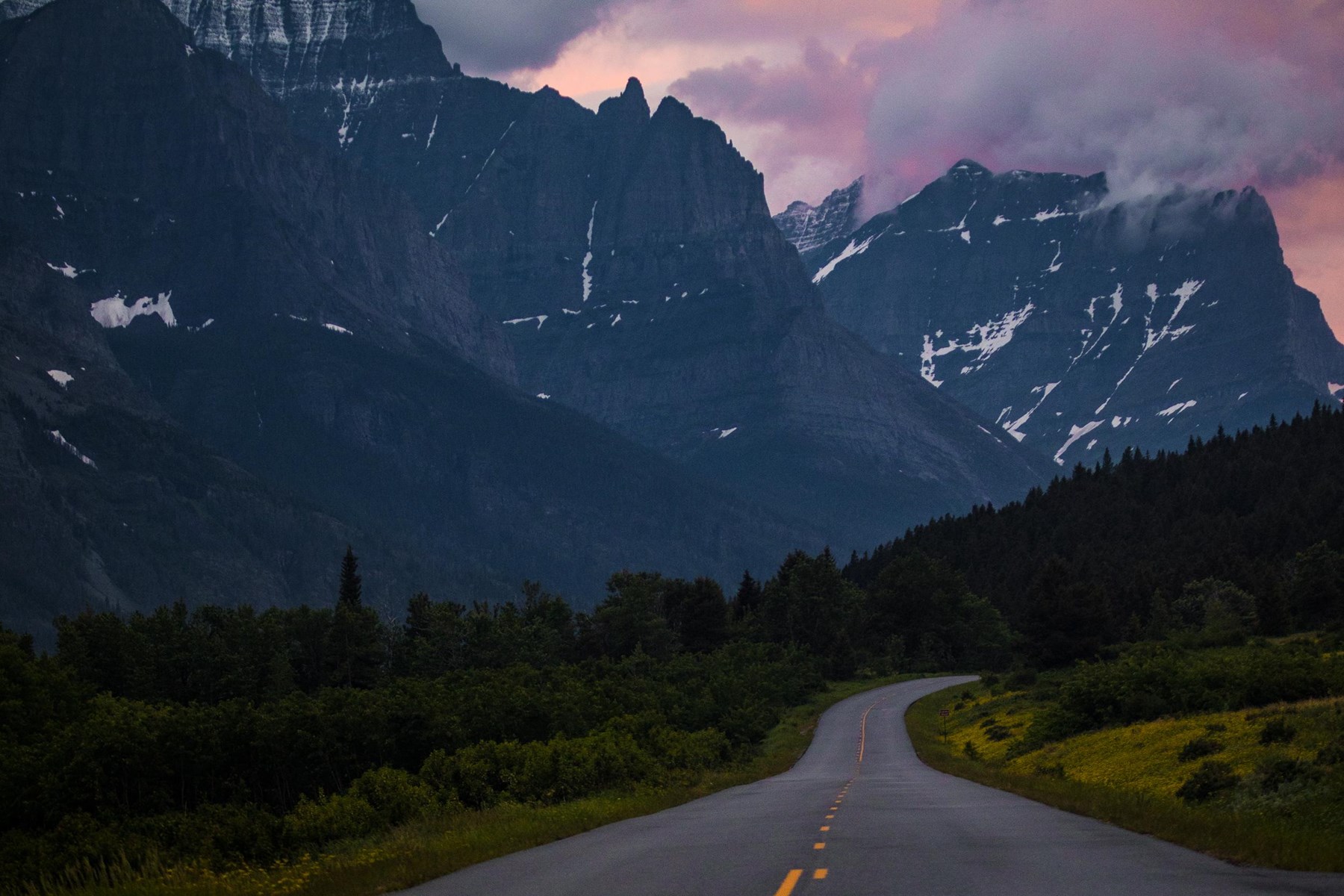 A winding road beneath dramatic mountain peaks at sunset in Glacier National Park, Montana